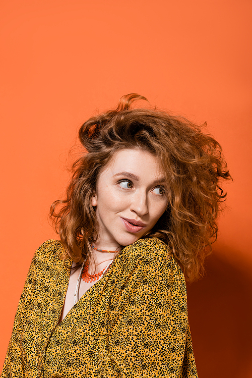 Astonished young red haired woman in yellow blouse with abstract pattern and necklaces looking away and standing on orange background, stylish casual outfit and summer vibes concept, Youth Culture