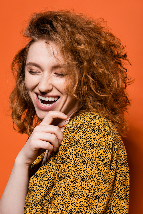 Portrait of young and positive redhead woman in yellow blouse with abstract pattern standing with closed eyes on orange background, stylish casual outfit and summer vibes concept, Youth Culture