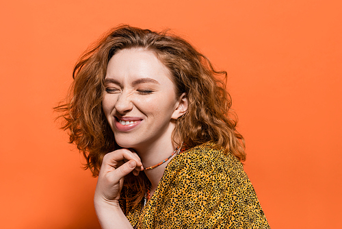 Joyful young redhead woman with closed eyes wearing yellow blouse with abstract print and touching necklaces on orange background, stylish casual outfit and summer vibes concept, Youth Culture