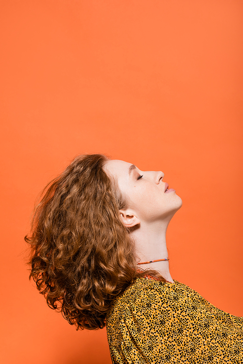 Side view of stylish young redhead woman with natural makeup in blouse with modern abstract print standing isolated on orange, stylish casual outfit and summer vibes concept, Youth Culture