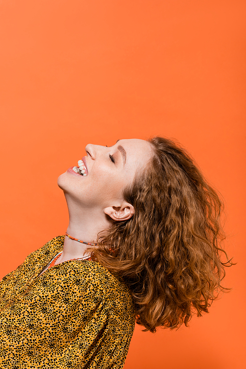 Smiling young red haired woman in necklaces and blouse with modern abstract print having fun and standing isolated on orange, stylish casual outfit and summer vibes concept, Youth Culture