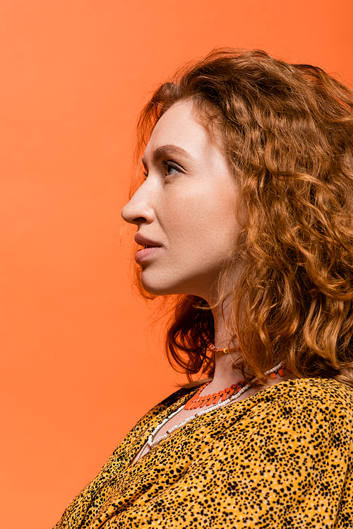 Side view of stylish redhead woman in necklaces and yellow blouse with abstract pattern looking away while standing isolated on orange, stylish casual outfit and summer vibes concept, Youth Culture