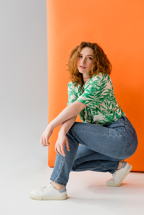 Full length of stylish redhead woman in blouse with floral pattern and jeans looking at camera while posing on orange and grey background, trendy casual summer outfit concept, Youth Culture