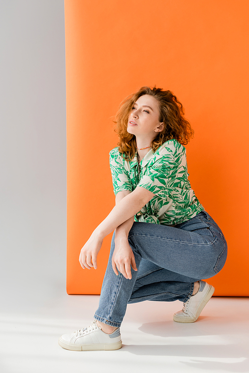 Full length of young red haired woman in blouse with floral pattern and jeans looking away while posing on orange and grey background, trendy casual summer outfit concept, Youth Culture