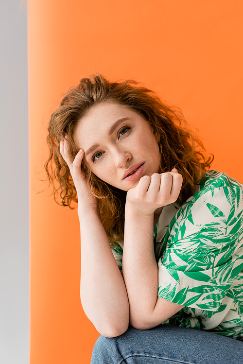 Portrait of young redhead woman in blouse with floral pattern and jeans looking at camera while standing on orange and grey background, trendy casual summer outfit concept, Youth Culture