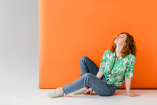 Relaxed young red haired woman with closed eyes in blouse with floral print and jeans sitting on grey and orange background, trendy casual summer outfit concept, Youth Culture
