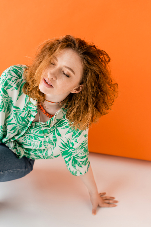 Stylish young redhead woman in blouse with floral pattern, necklaces and jeans posing with closed eyes on orange background, trendy casual summer outfit concept, Youth Culture