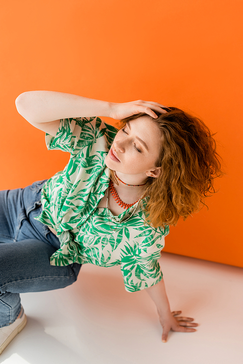Stylish young red haired woman in jeans and blouse with floral pattern touching head and closing eyes while posing on orange background, trendy casual summer outfit concept, Youth Culture