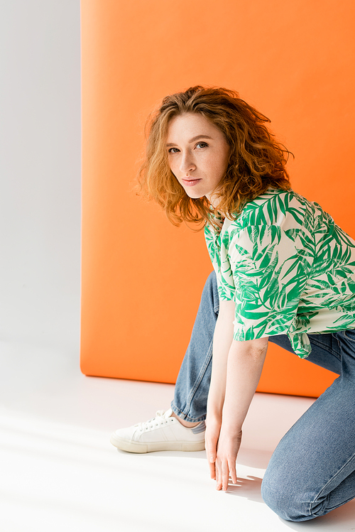 Stylish redhead woman with natural makeup wearing blouse with floral print and jeans while looking at camera on orange background, trendy casual summer outfit concept, Youth Culture
