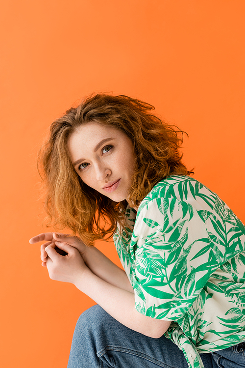 Portrait of stylish and confident redhead model in blouse with floral pattern and jeans looking at camera isolated on orange, trendy casual summer outfit concept, Youth Culture