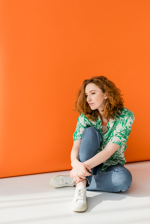 Full length of young red haired woman in modern blouse with floral pattern and jeans sitting on grey and orange background, trendy casual summer outfit concept, Youth Culture
