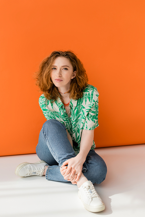 Full length of young red haired woman in modern blouse with floral pattern and jeans looking at camera while sitting on orange background, trendy casual summer outfit concept, Youth Culture