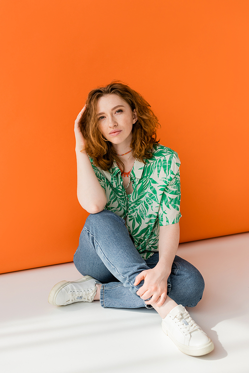 Full length of trendy young redhead woman in blouse with floral pattern, jeans and necklaces looking at camera and sitting on orange background, trendy casual summer outfit concept, Youth Culture