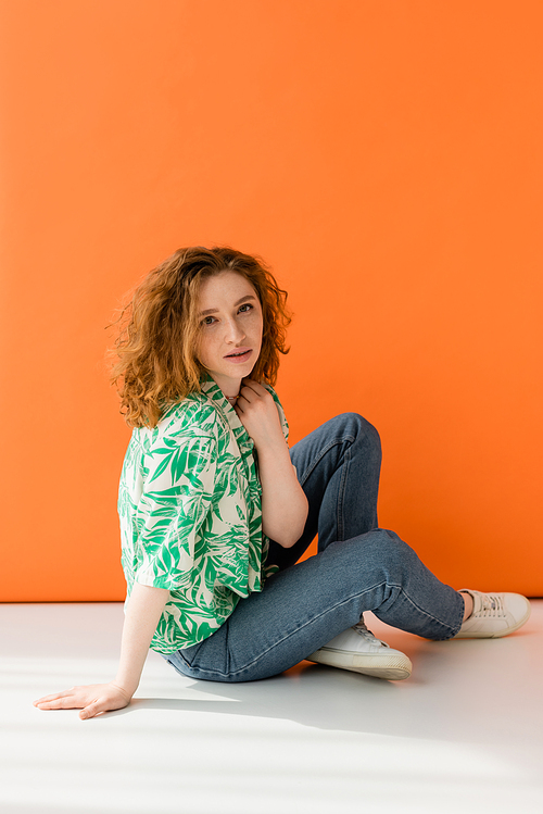 Young red haired model in modern blouse with floral pattern and jeans looking at camera while sitting on orange background, trendy casual summer outfit concept, Youth Culture