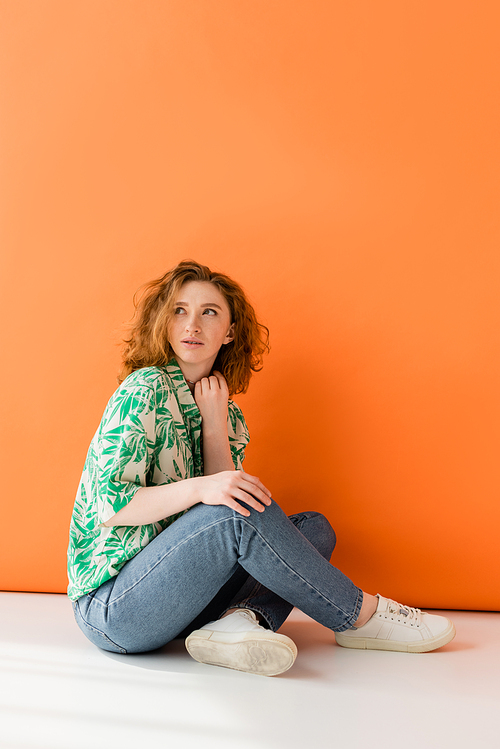 Full length of young redhead woman in stylish blouse with floral pattern and jeans looking away while sitting on orange background, trendy casual summer outfit concept, Youth Culture