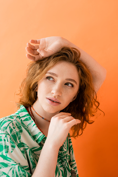 Portrait of dreamy and stylish redhead woman in modern blouse with floral pattern looking away and posing isolated on orange, trendy casual summer outfit concept, Youth Culture