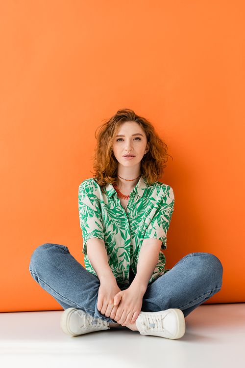 Full length of young red haired woman with natural makeup, jeans and blouse with floral pattern looking at camera on orange background, trendy casual summer outfit concept, Youth Culture