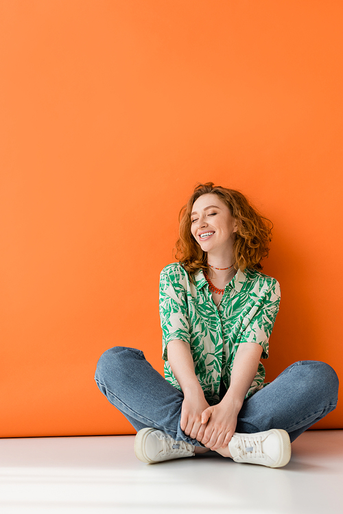 Full length of positive young redhead woman in blouse with floral pattern and jeans sitting with closed eyes on orange background, trendy casual summer outfit concept, Youth Culture