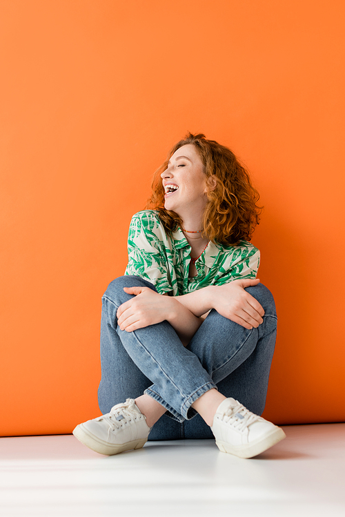 Joyful young red haired model in stylish blouse with floral pattern and modern jeans touching knees and sitting on orange background, trendy casual summer outfit concept, Youth Culture