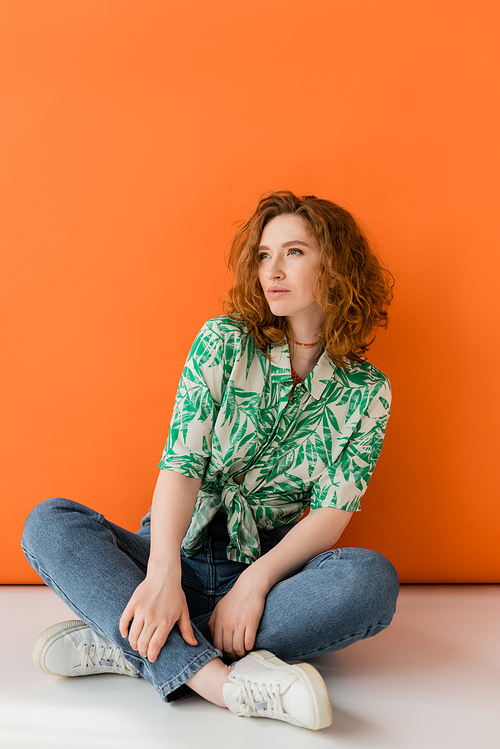 Confident young redhead woman in modern blouse with floral pattern and jeans looking away while sitting on orange background, trendy casual summer outfit concept, Youth Culture