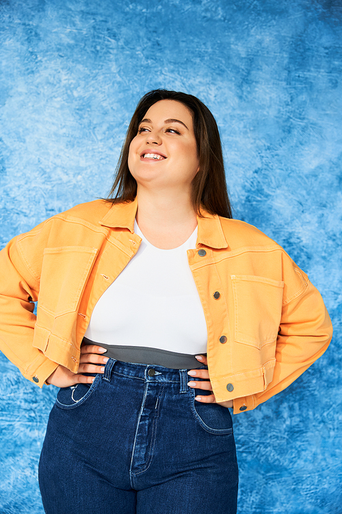 cheerful plus size woman with long hair and natural makeup wearing crop top, orange jacket and denim jeans while posing with hands on hips and looking away on mottled blue background, body positive