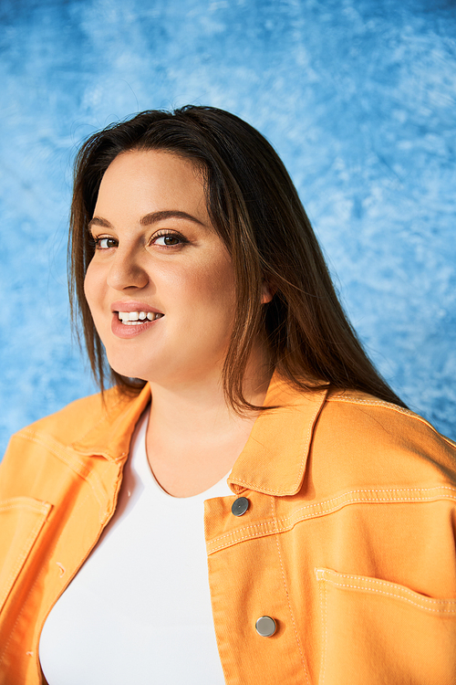portrait of positive plus size woman with long hair and natural makeup wearing crop top and orange jacket while posing and looking at camera on mottled blue background, body positive