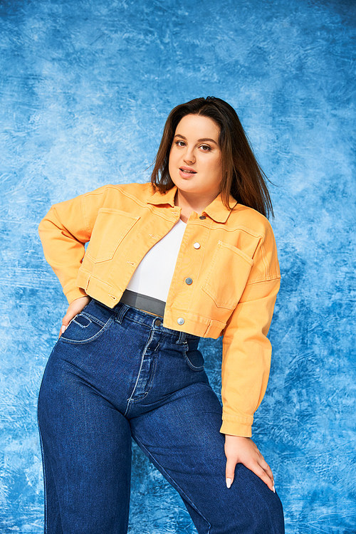 woman with plus size body and long hair, wearing crop top, orange jacket and denim jeans while posing with hand on hip and looking at camera on mottled blue background, body positive