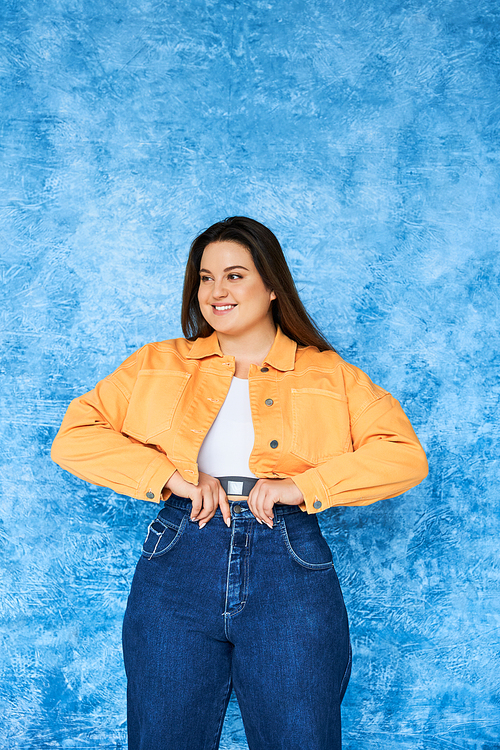 positive plus size woman with long hair and natural makeup wearing crop top, orange jacket and adjusting denim jeans while posing and looking away on mottled blue background, body positive