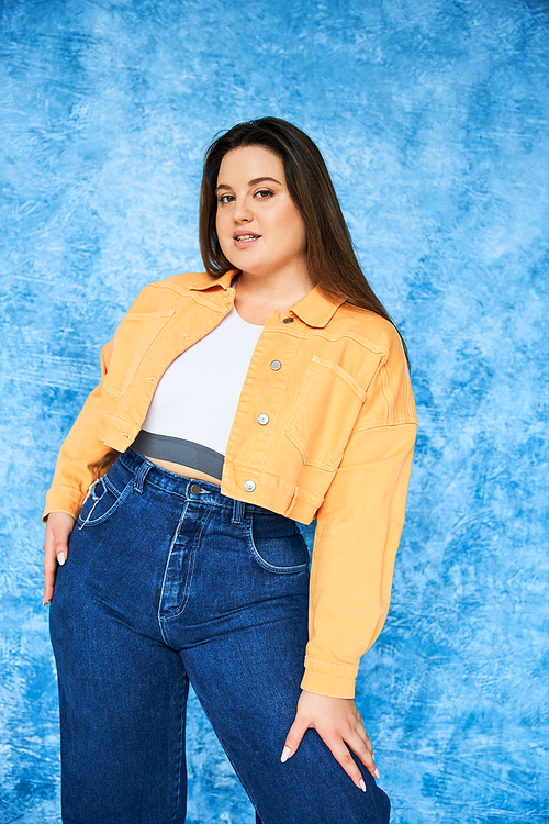 plus size woman with brunette long hair and natural makeup wearing crop top, orange jacket and denim jeans while posing and looking at camera on mottled blue background, body positive