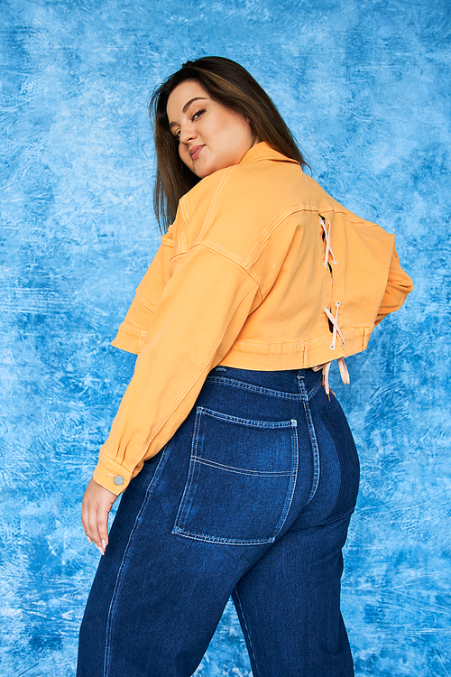 alluring plus size woman with long hair and natural makeup wearing crop top, orange jacket and denim jeans while posing and looking at camera on mottled blue background, body positive