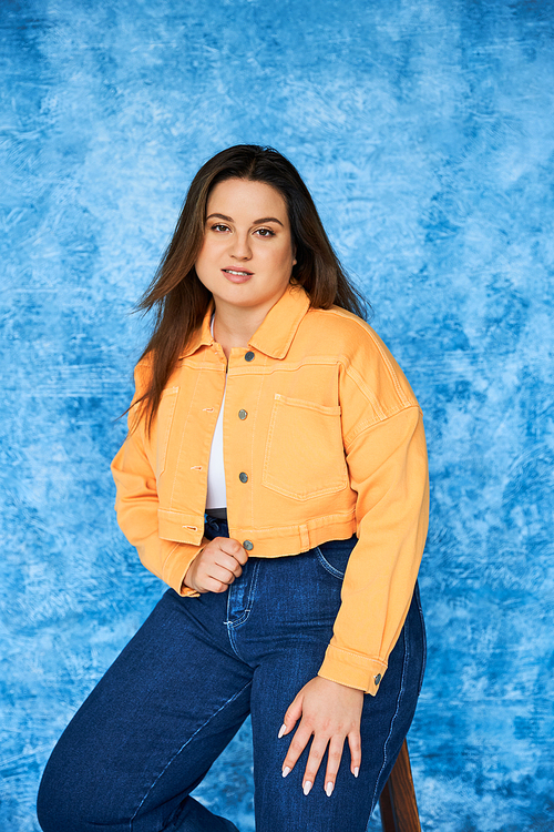 brunette plus size woman with long hair and natural makeup wearing orange jacket and denim jeans while sitting on stool and looking at camera on mottled blue background, body positive