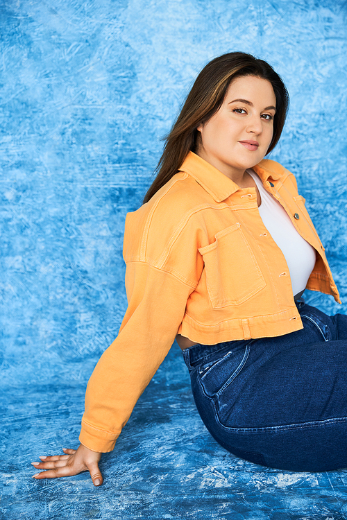 body positive and pretty plus size woman with long hair and natural makeup wearing crop top, orange jacket and denim jeans while sitting and looking at camera on mottled blue background