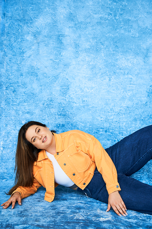 plus size woman with brunette hair and natural makeup wearing crop top, orange jacket and denim jeans while posing and looking at camera on mottled blue background, body positive