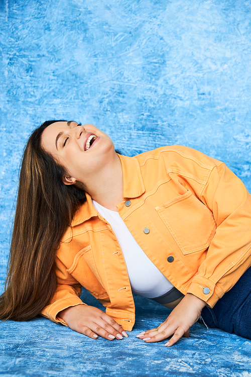 body positive and happy plus size woman with long hair and natural makeup laughing with closed eyes while posing in orange jacket and denim jeans on mottled blue background