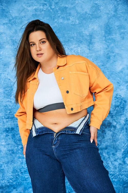 body positive and brunette plus size woman with long hair and natural makeup posing in crop top, orange jacket and denim jeans while standing and looking at camera on mottled blue background