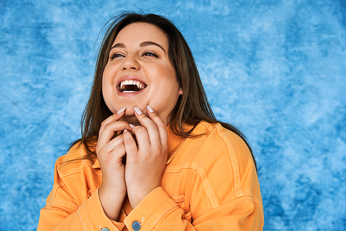 portrait of body positive and happy plus size woman with brunette hair and natural makeup laughing while touching face and posing in orange jacket on mottled blue background