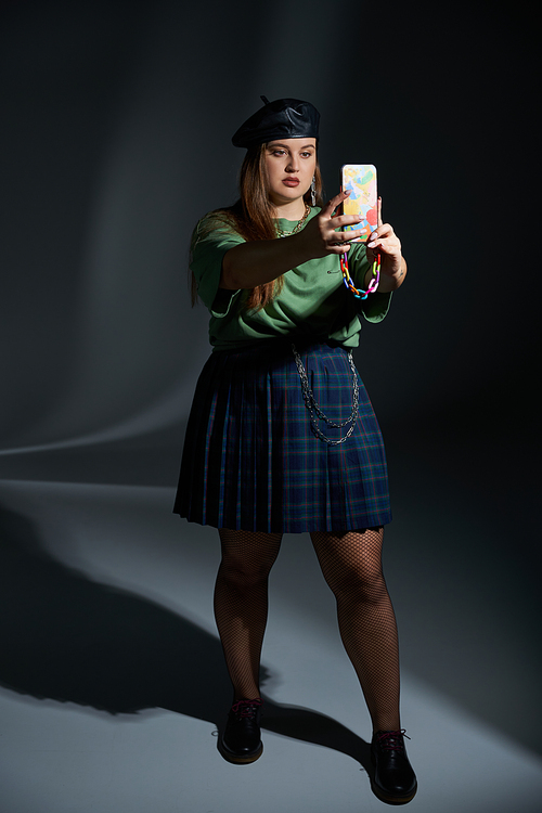 plus size woman posing in leather beret, green t-shirt, plaid skirt with chains, fishnet tights and black shoes, taking selfie on smartphone on dark background, tattoo translation: harmony