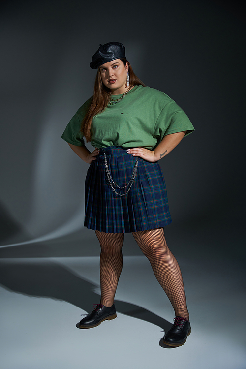 full length of plus size woman posing in leather beret, green t-shirt, plaid skirt with chains and fishnet tights looking at camera and posing on dark background, tattoo translation: harmony