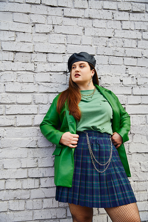 chic plus size woman posing in plaid skirt with chains, green leather jacket, black beret and fishnet tights while looking away and standing near brick wall on urban street, body positive