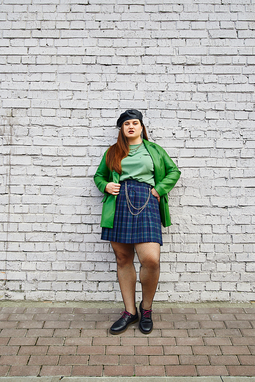 full length of brunette plus size woman posing in leather jacket with beret, plaid skirt with chains, fishnet tights and black shoes while standing near brick wall on urban street