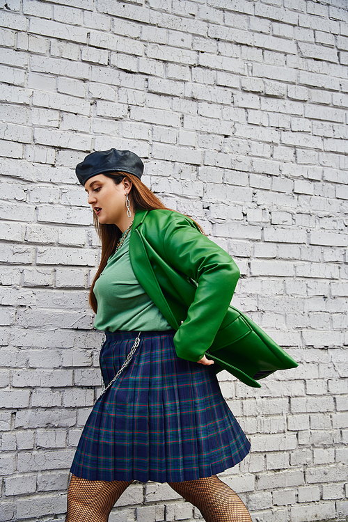 side view of stylish plus size woman in green leather jacket, black beret, plaid skirt with chains and fishnet tights walking near brick wall on urban street, body positive, self-love, urban chic
