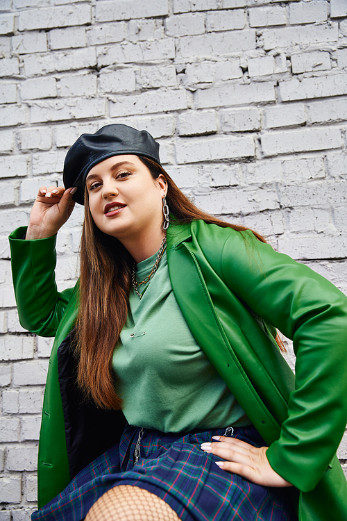 stylish plus size woman in green leather jacket smiling while touching black beret and posing in plaid skirt and fishnet tights near brick wall on urban street, body positive, self-love, urban chic