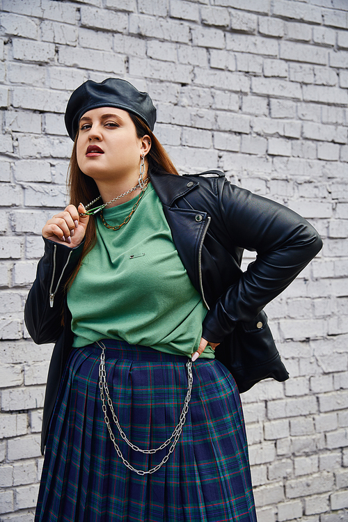 stylish plus size woman in leather jacket and black beret pulling chain necklace while looking at camera and standing with hand on hip near brick wall on urban street, body positive, urban chic