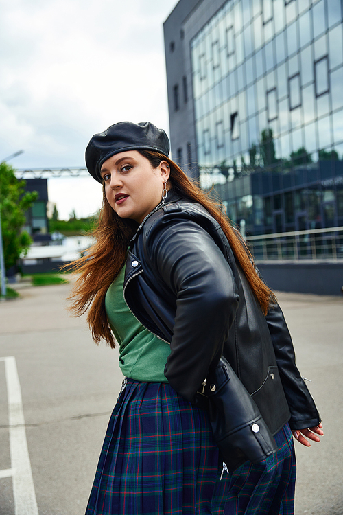 stylish woman with plus size body walking in leather jacket with black beret, plaid skirt and greet t-shirt near blurred modern building on urban street outdoors, body positive, looking at camera
