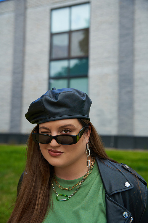 portrait of plus size brunette woman posing in stylish sunglasses, leather jacket with black beret and greet t-shirt winking near blurred modern building on urban street outdoors, body positive