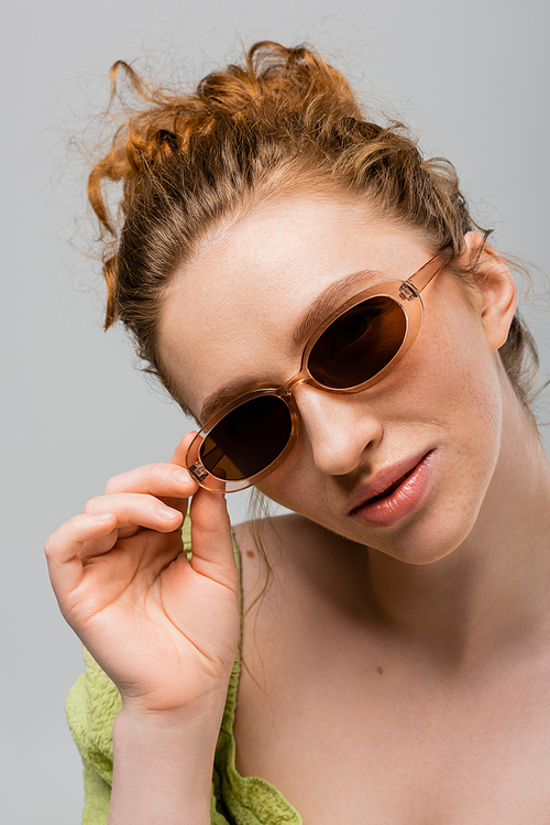 Portrait of confident, red haired and freckled woman in green blouse touching sunglasses while posing and standing isolated on grey, trendy sun protection concept, Youth Culture
