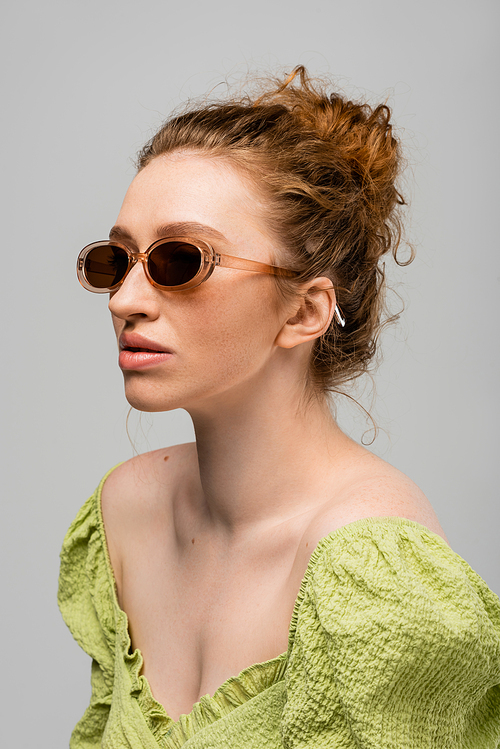 Portrait of stylish young red haired woman in green blouse and modern sunglasses posing while standing isolated on grey background, trendy sun protection concept, fashion model