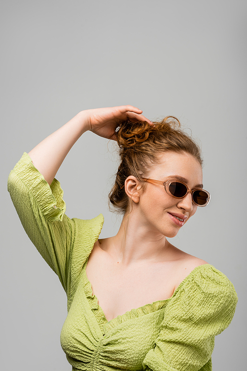 Smiling young redhead woman in summer green blouse and sunglasses touching hair while standing isolated on grey background, trendy sun protection concept, fashion model