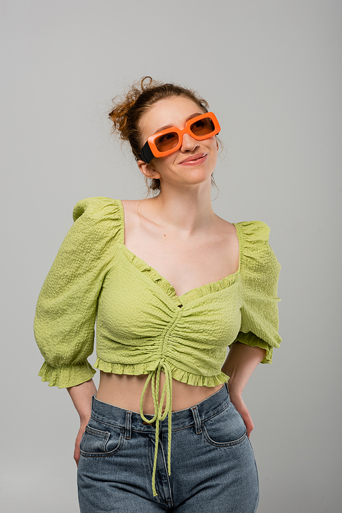 Smiling young woman in green blouse, jeans and sunglasses posing and looking away while standing isolated on grey background, fashion model, trendy sun protection concept