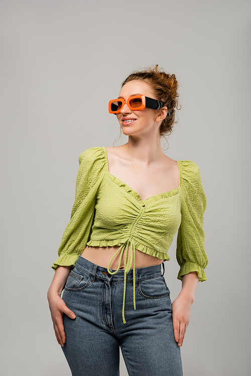 Joyful redhead woman in jeans, green blouse and sunglasses posing and looking away while standing isolated on grey background, trendy sun protection concept, fashion model
